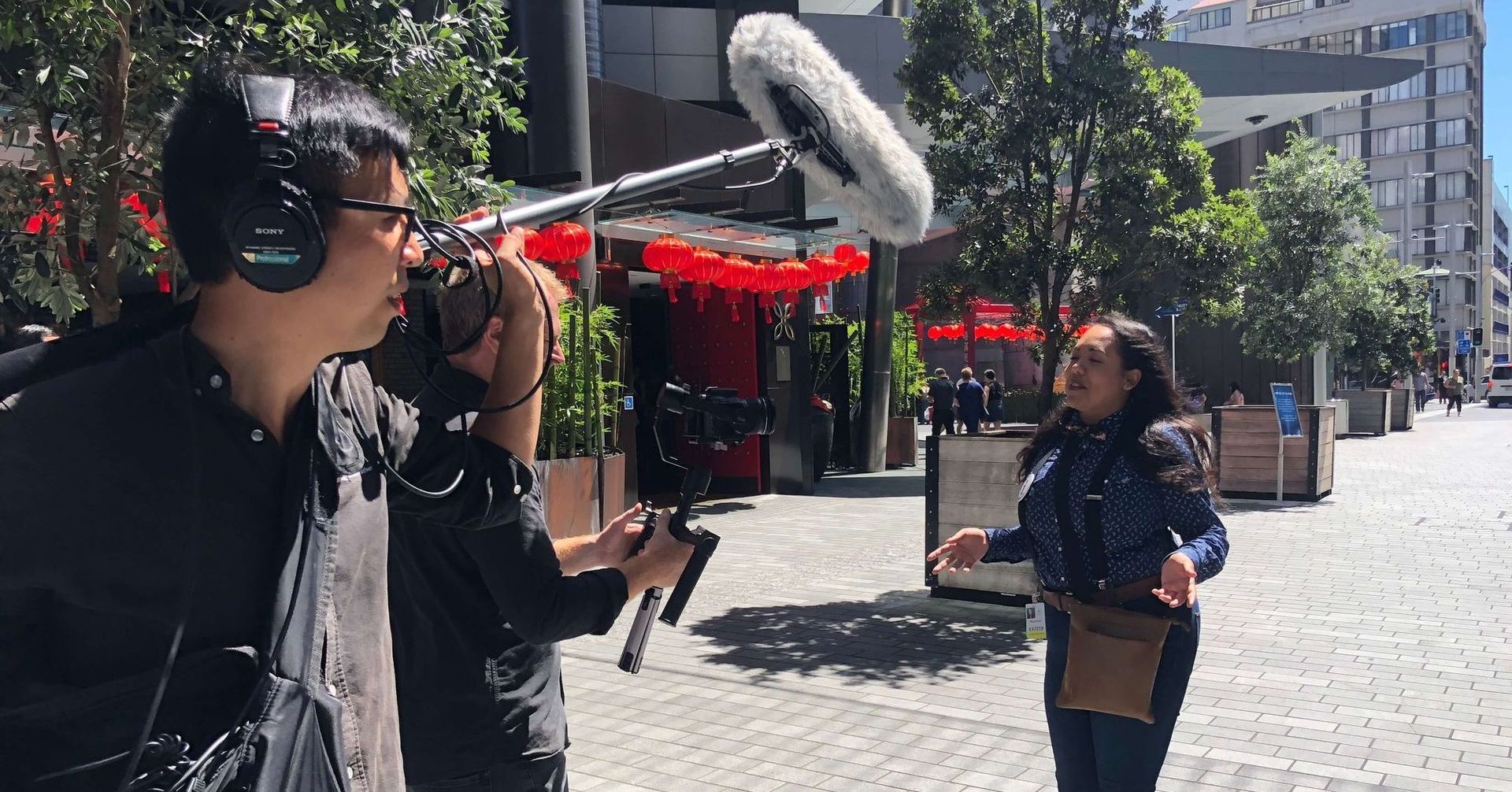 A film crew records a woman outside a restaurant. A boom mic, camera, and operator are visible.