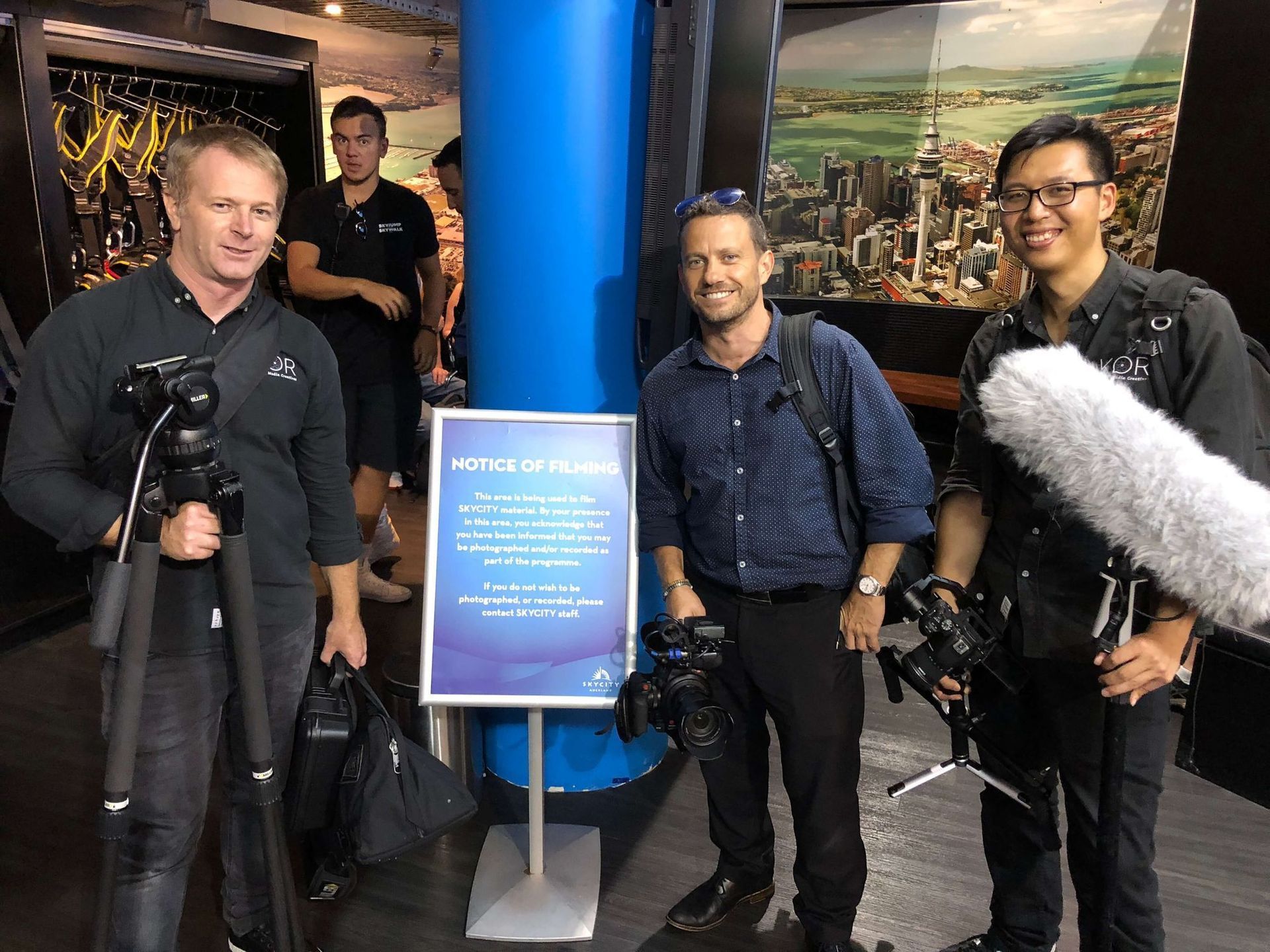 Three people with cameras posing near a sign and a blue pillar in a building, likely filming.