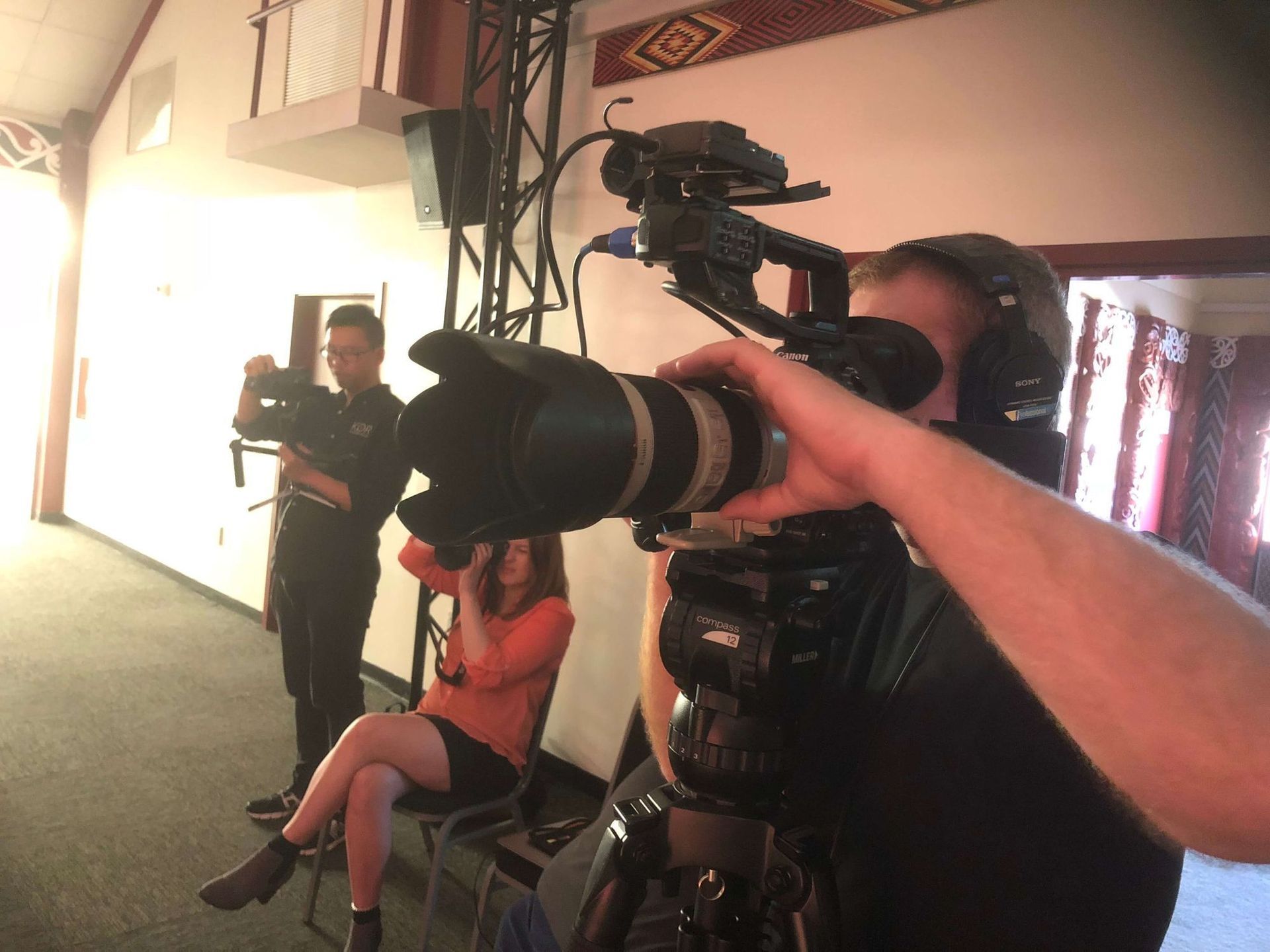 People filming. One cameraman is close-up, aiming the camera. Others are in the background, in a room with light.