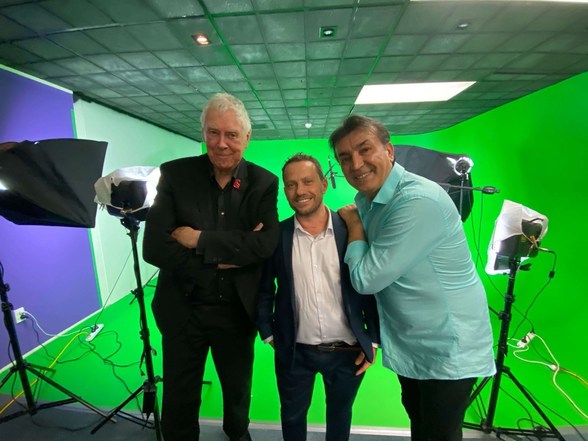 Three men pose in a studio with green and blue screens. Two men are smiling, and one has his arm around the middle man.