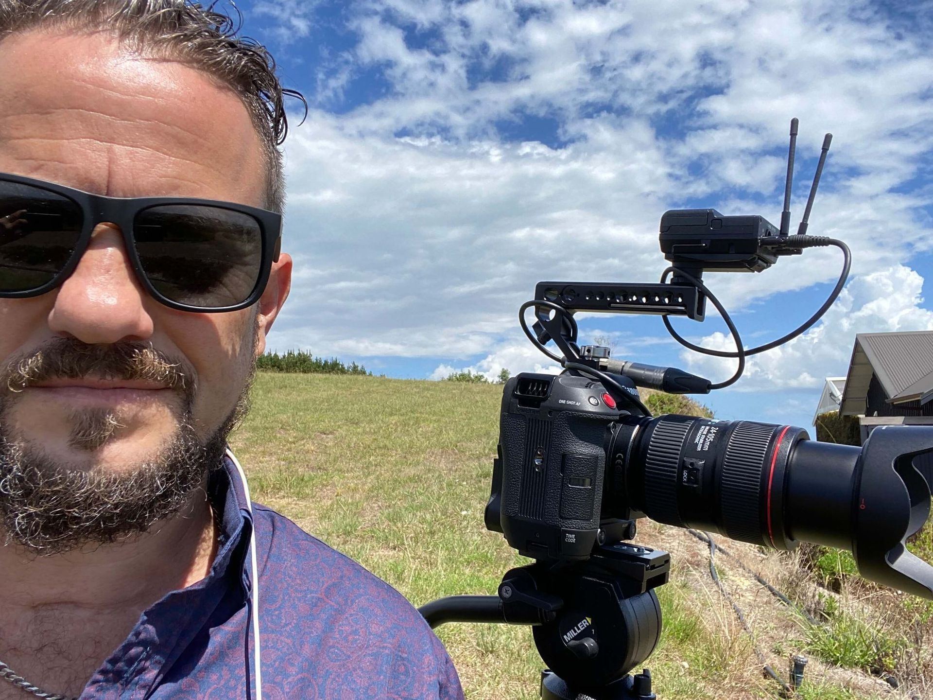 Craig Walsh in sunglasses films with a camera on a tripod outdoors, cloudy sky in background.