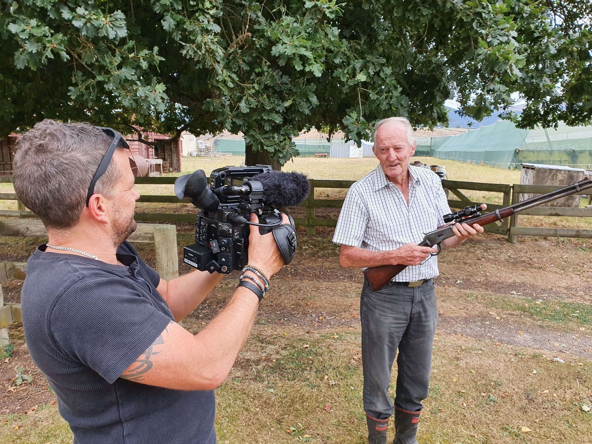 Man filming another man holding a rifle outdoors.