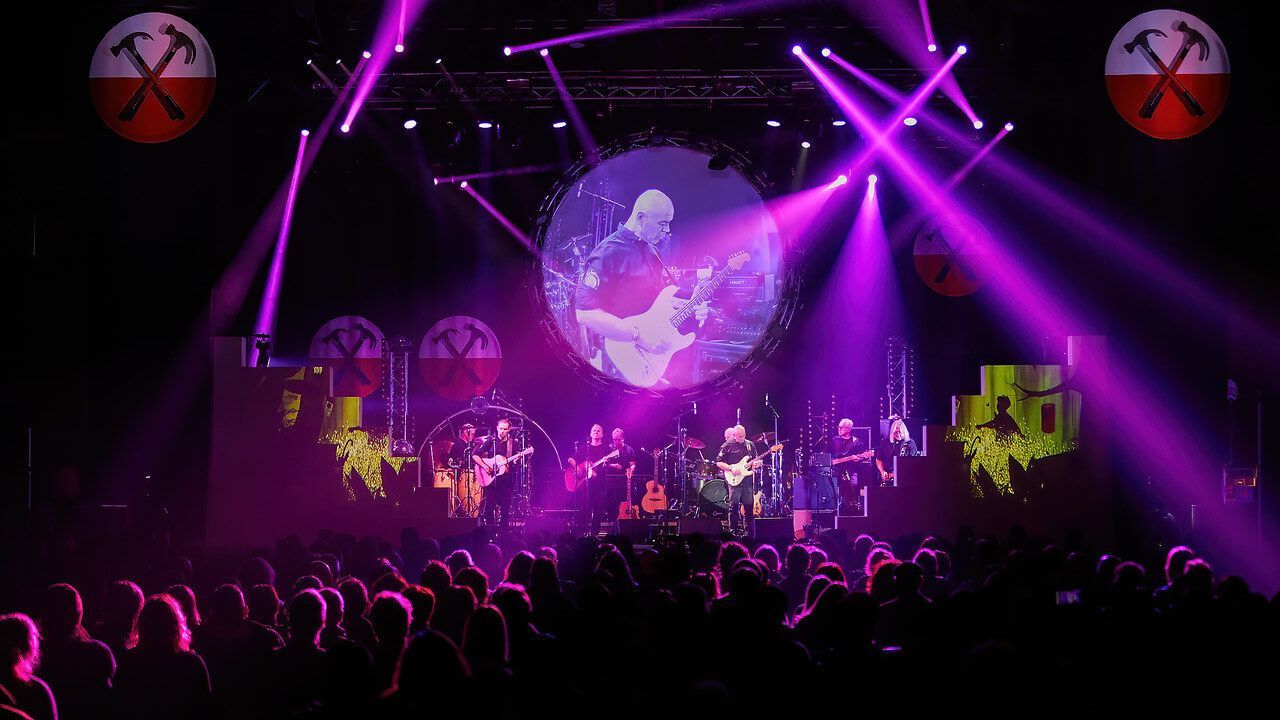 Pink Floyd Experience concert performance under bright purple lights, audience silhouetted below. Giant circular projection.