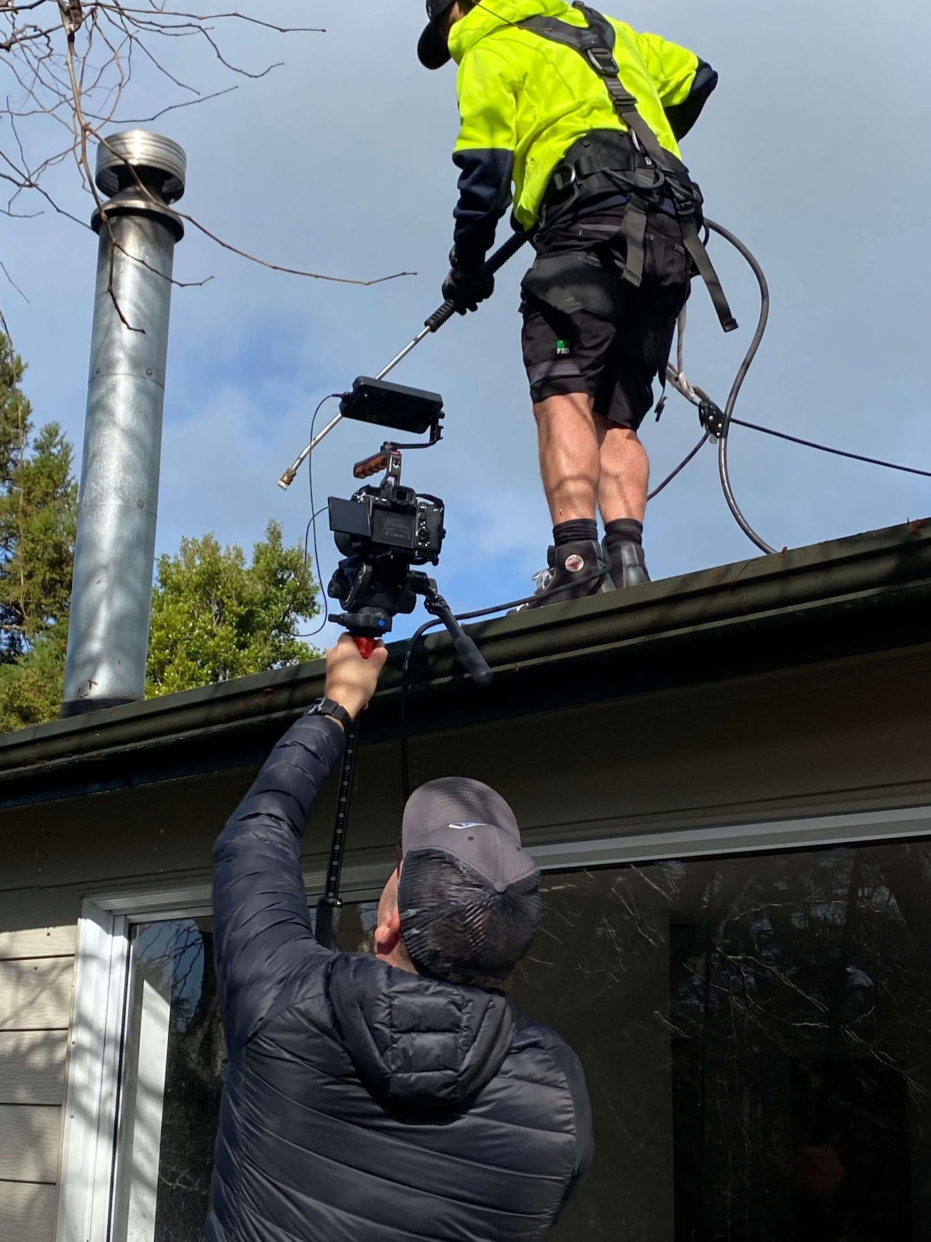 Two people working on a roof for Monster Wash. One cleans a chimney, the other films with a camera.