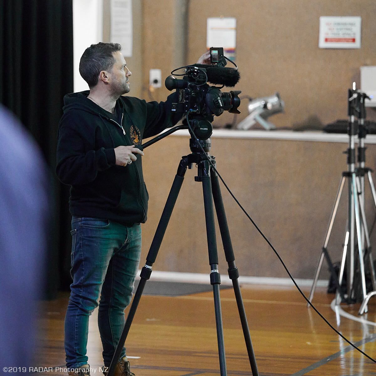 Man operating video camera on tripod in a wood-floored room. Another tripod is in the background.