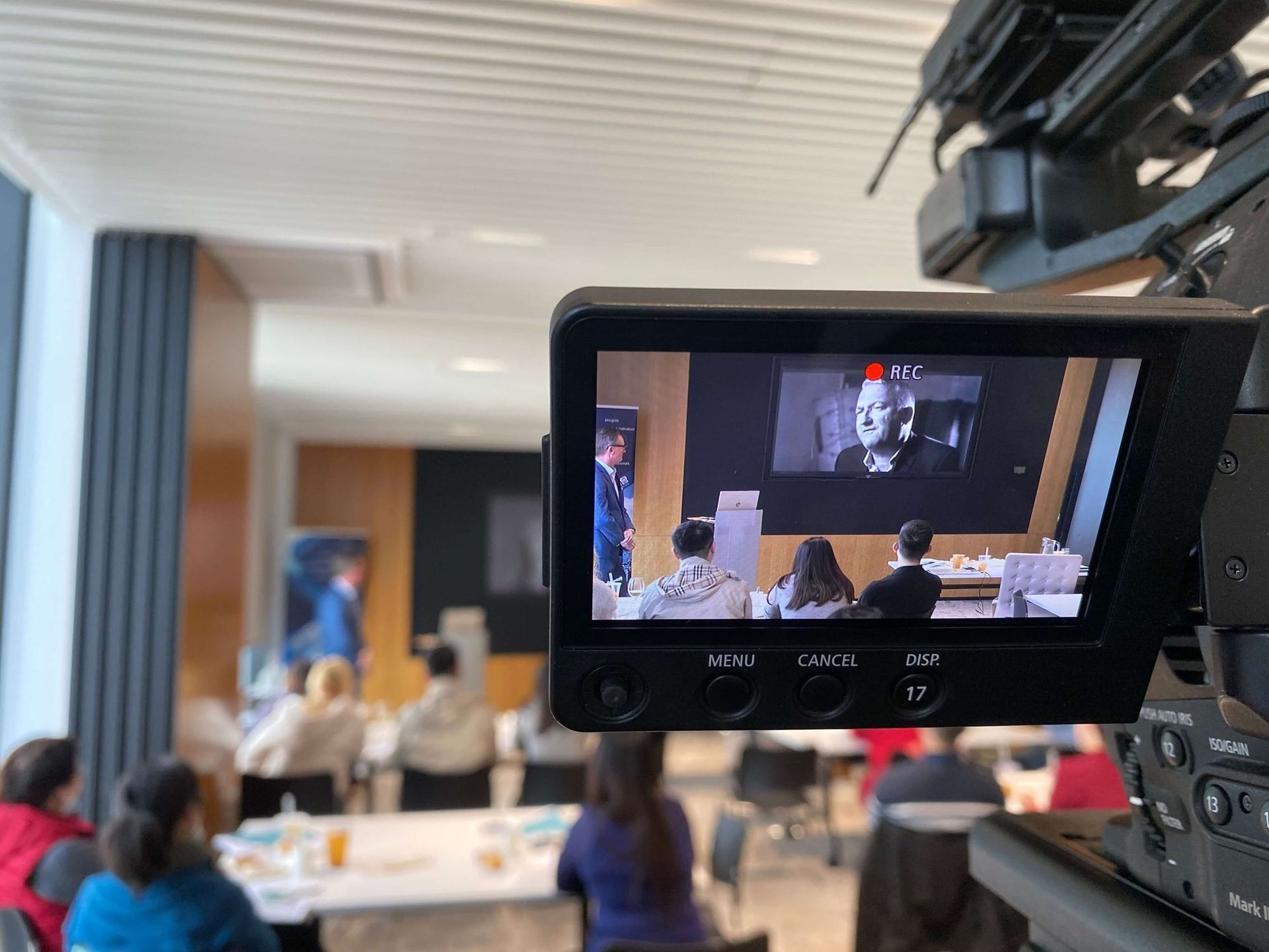 Camera screen shows a conference with a speaker on a screen. People sit at tables in a modern room.