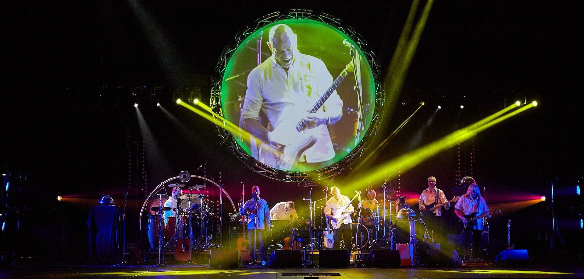 A band performs on stage. A large circular screen displays a musician. Yellow spotlights shine.