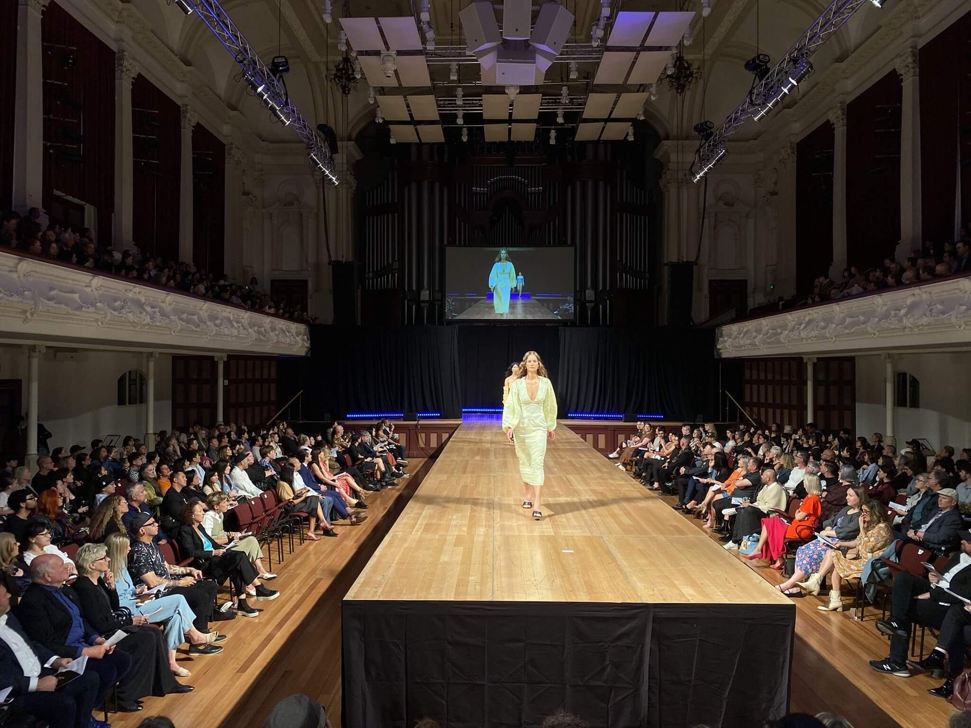 A fashion model in a light green outfit walks a runway at a show; audience seated on both sides.