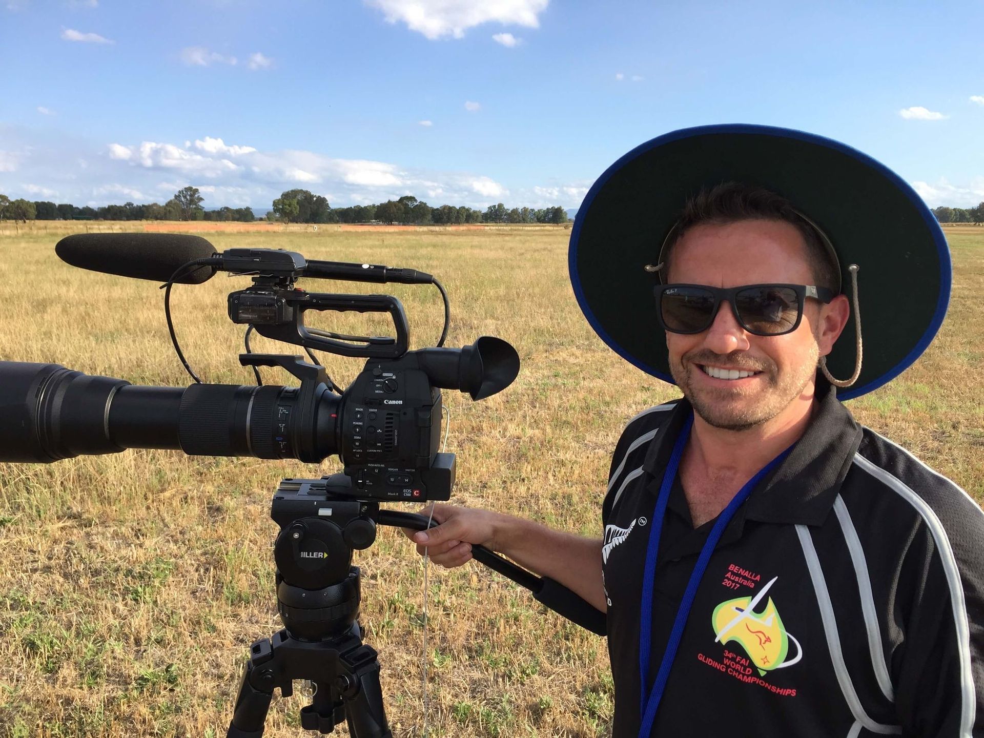 Craig Walsh in sunglasses and hat smiles next to a video camera on a tripod in a field.