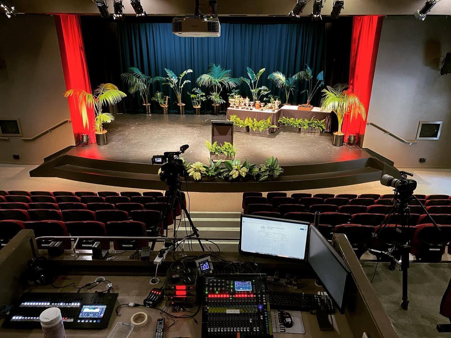 A stage set for a presentation with red curtains, greenery, and audio/video equipment in the foreground.