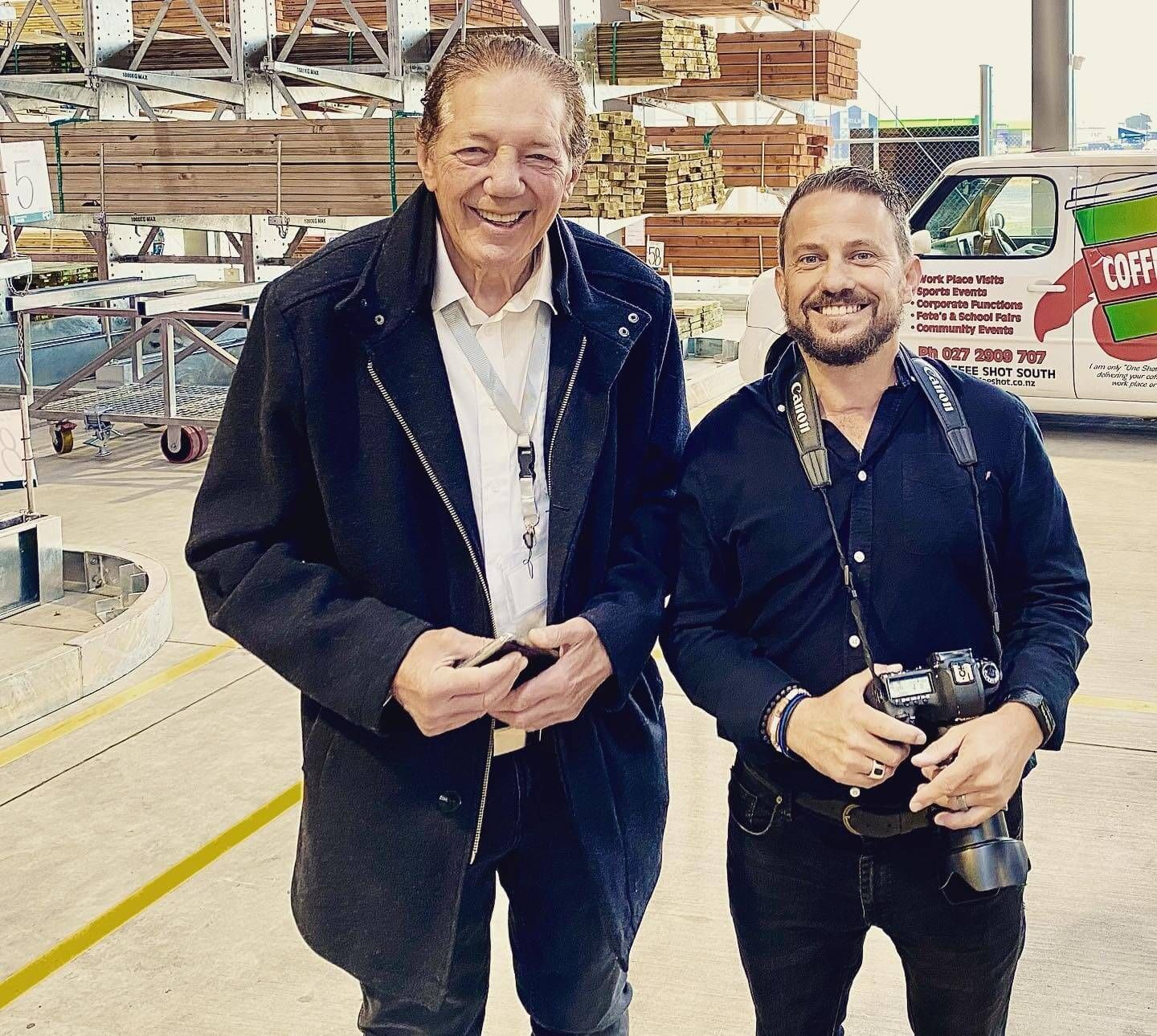 Two men smiling, Craig Walsh holding a camera with Tim Shadbolt in a lumberyard setting near a delivery van.