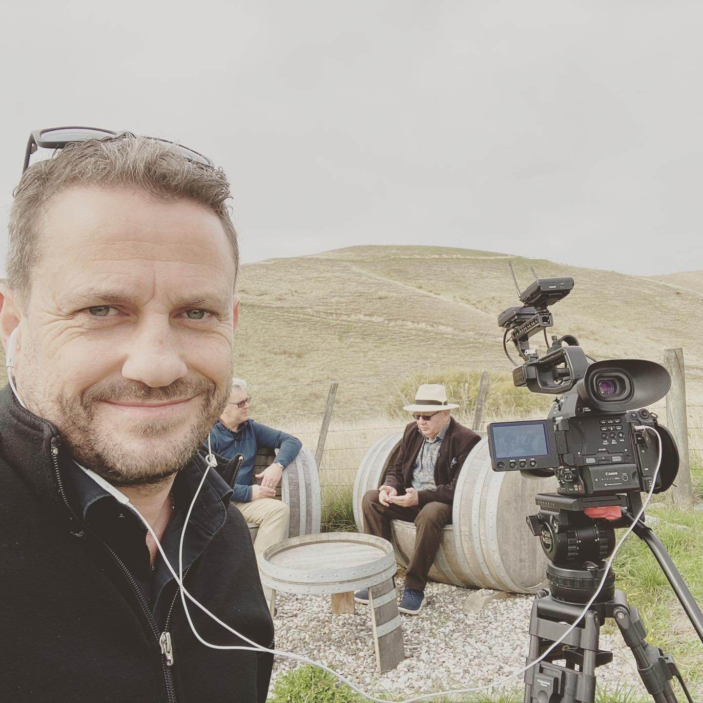 Craig Walsh with earbuds in selfie, filming two men in a vineyard setting. Camera on tripod.