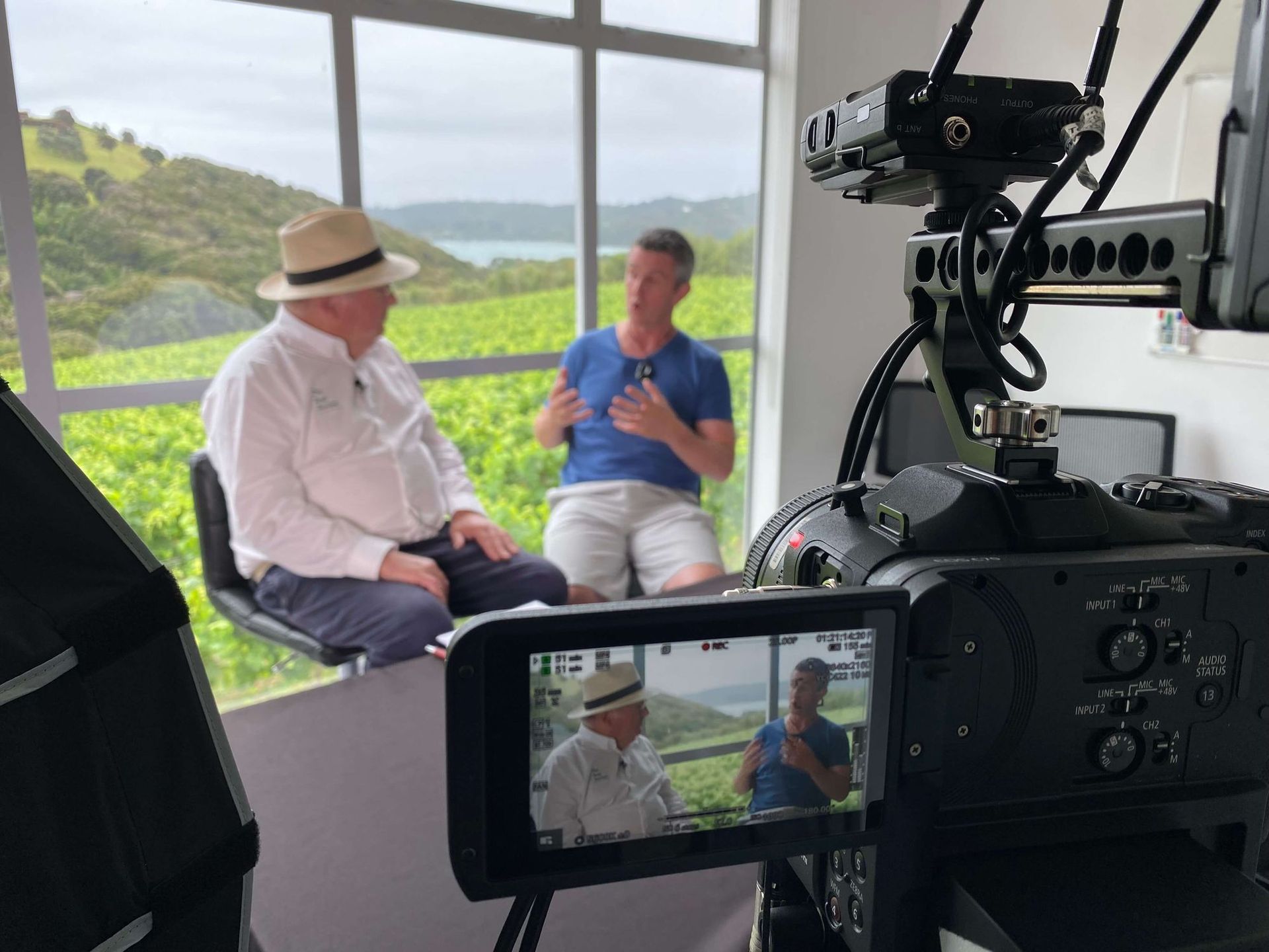 Two men in an interview, one wearing a hat, filmed against a vineyard backdrop.