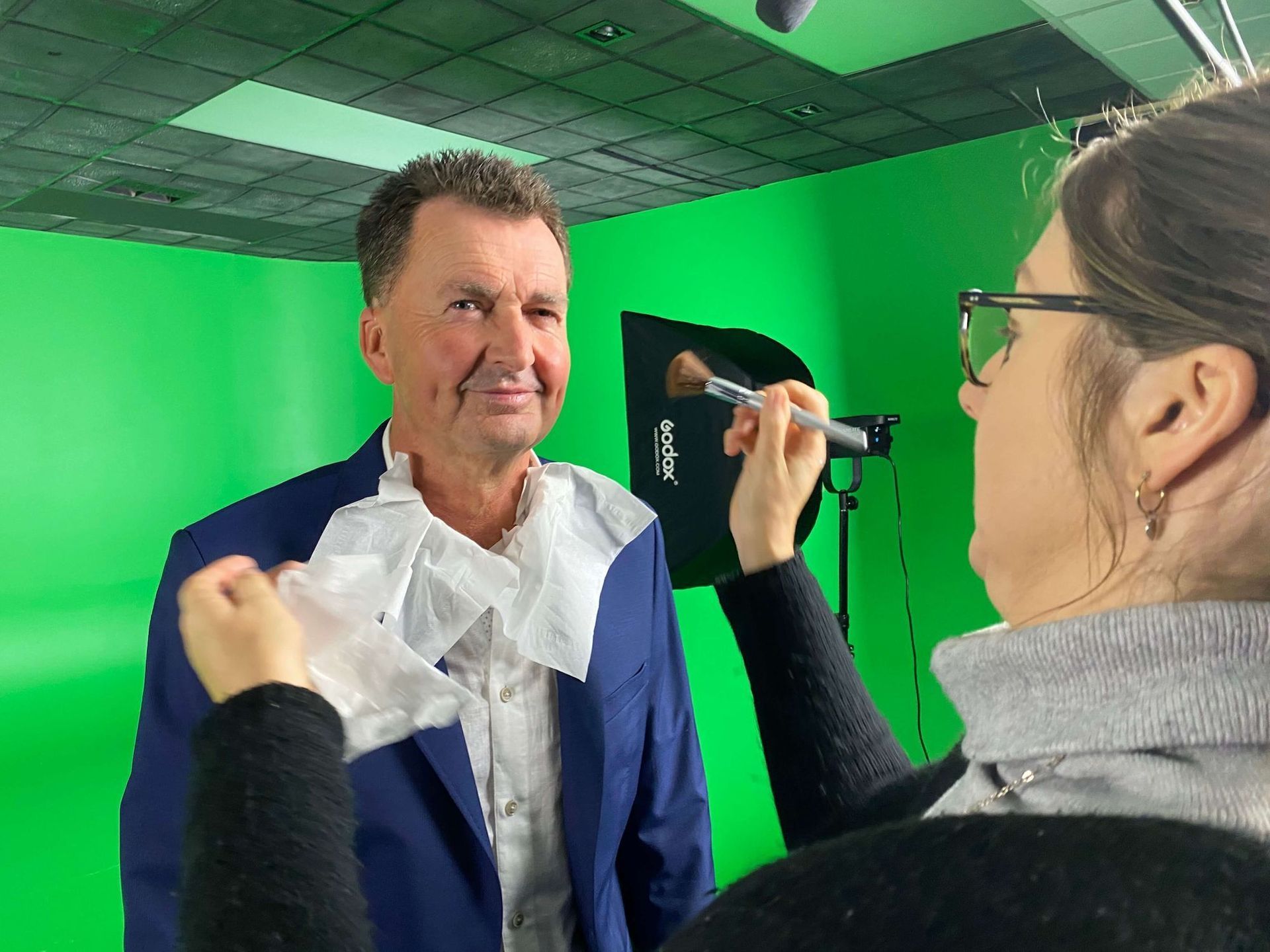 Makeup artist applying makeup to a man in front of a green screen, filming a video.