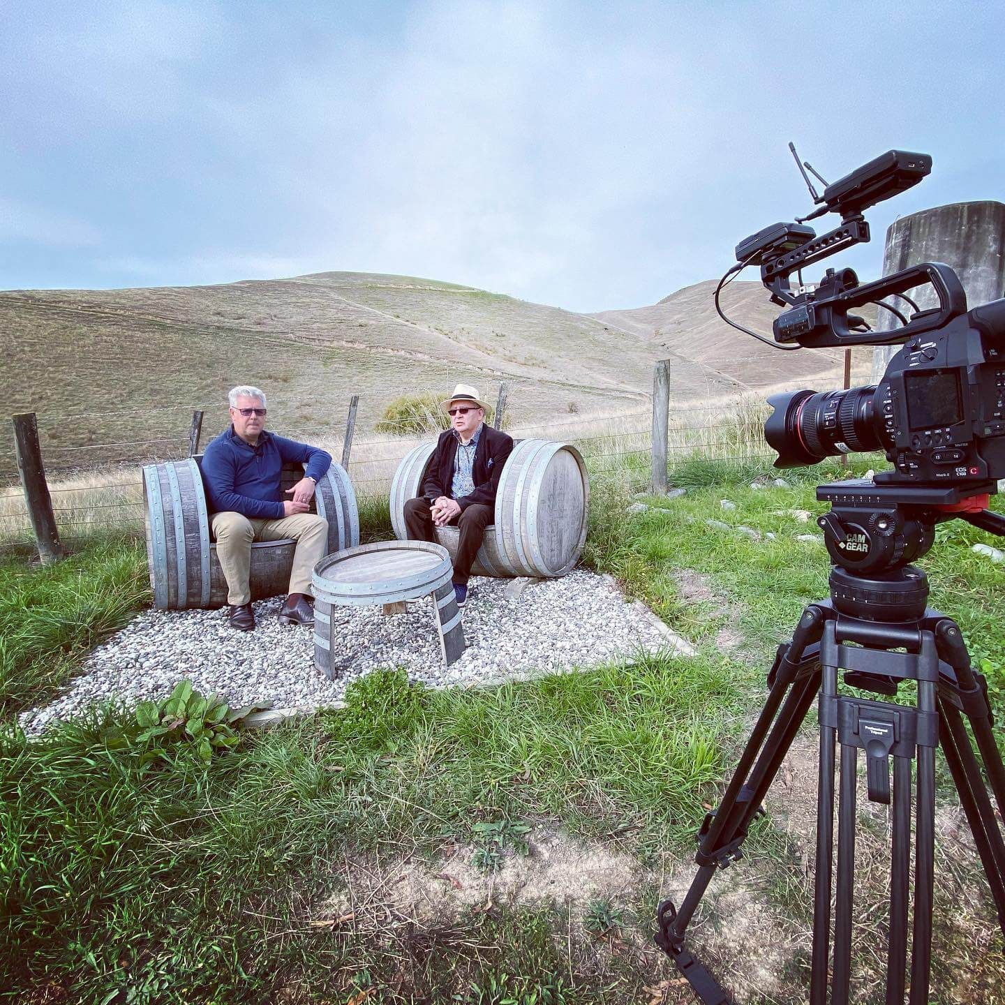 Two men seated on barrels, being filmed outdoors by a camera on a tripod, in a rural setting.