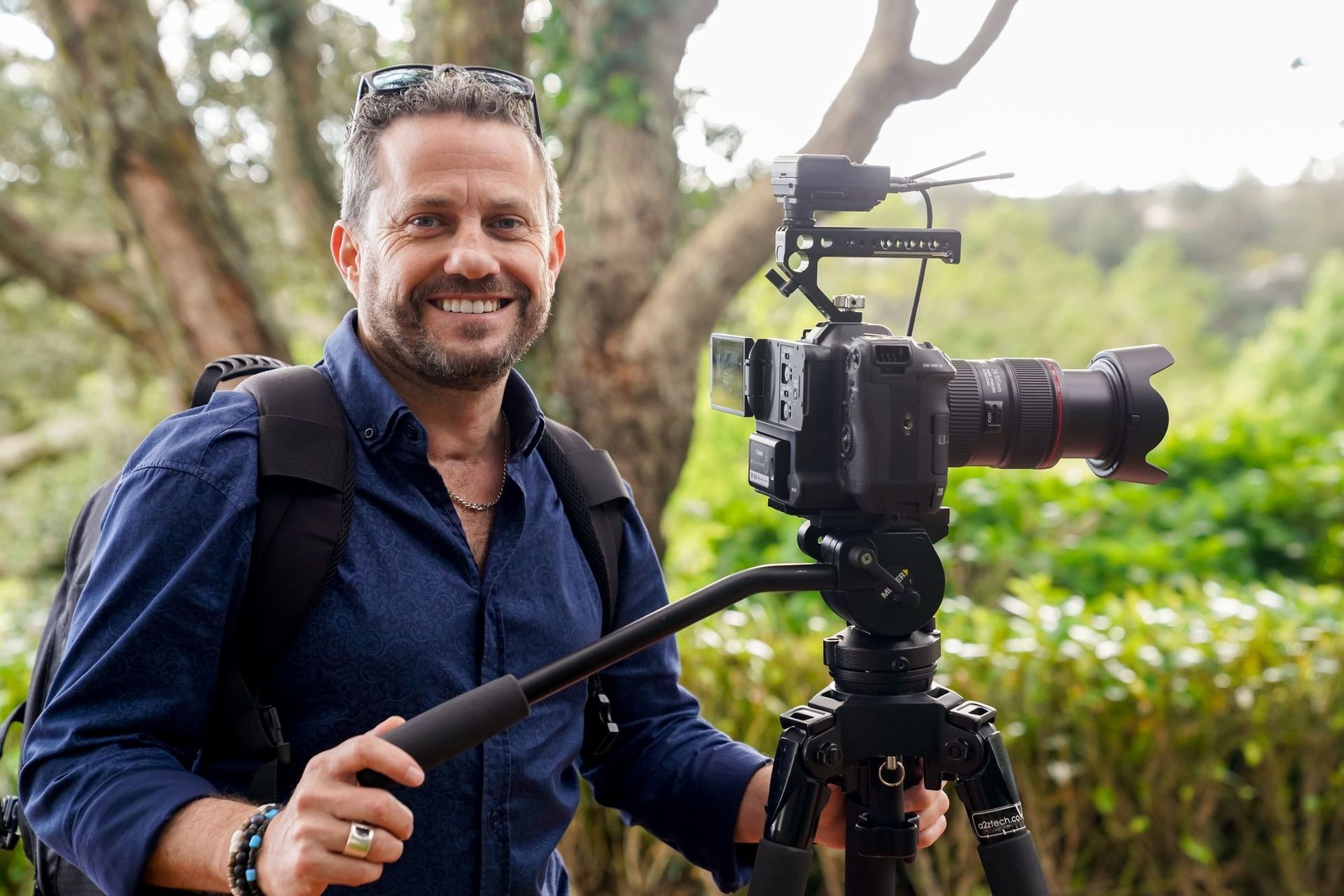 Craig Walsh with camera on tripod outdoors, wearing blue shirt and sunglasses.