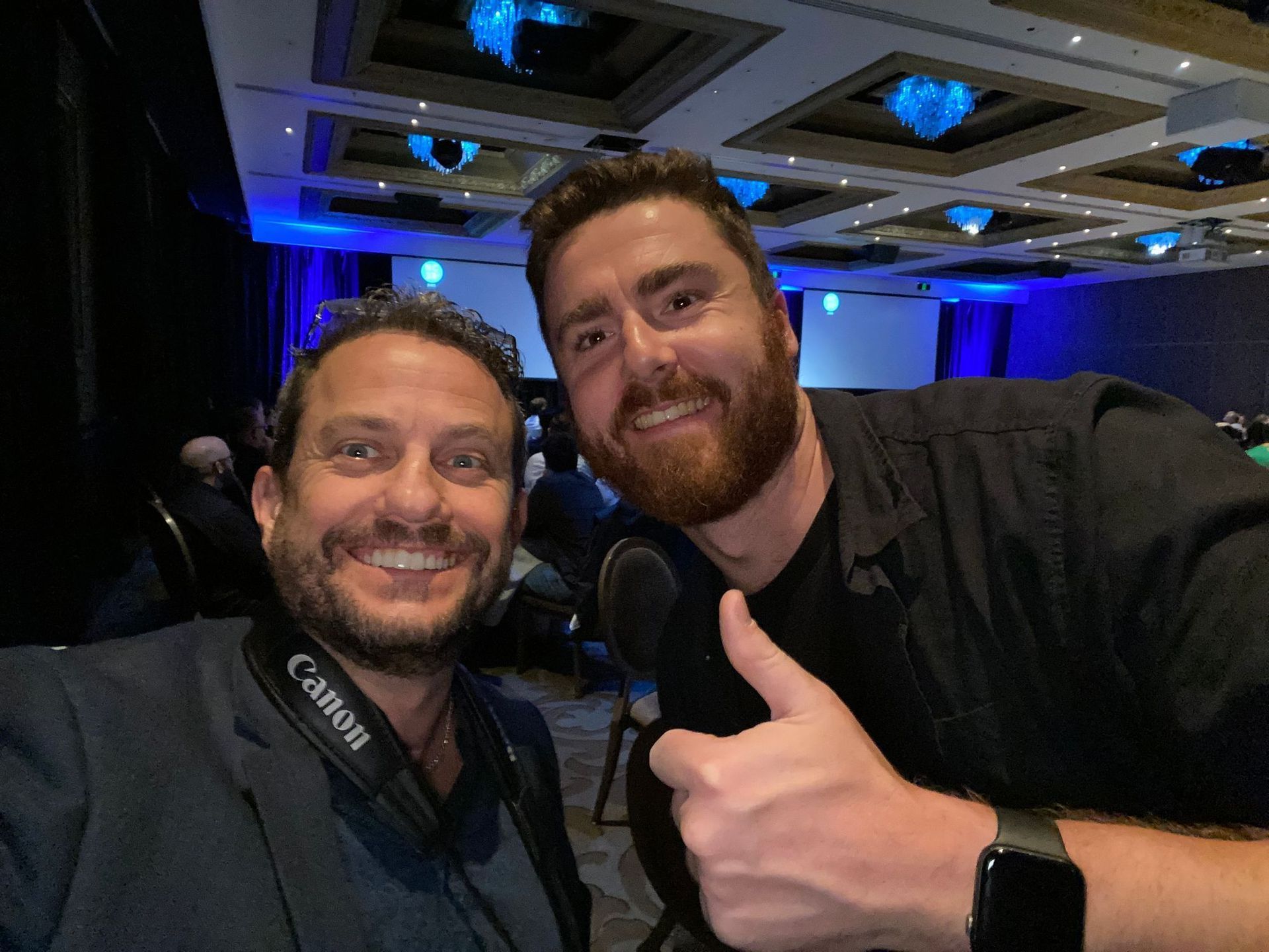 Two men smiling, taking a selfie in a conference room. One gives a thumbs-up.