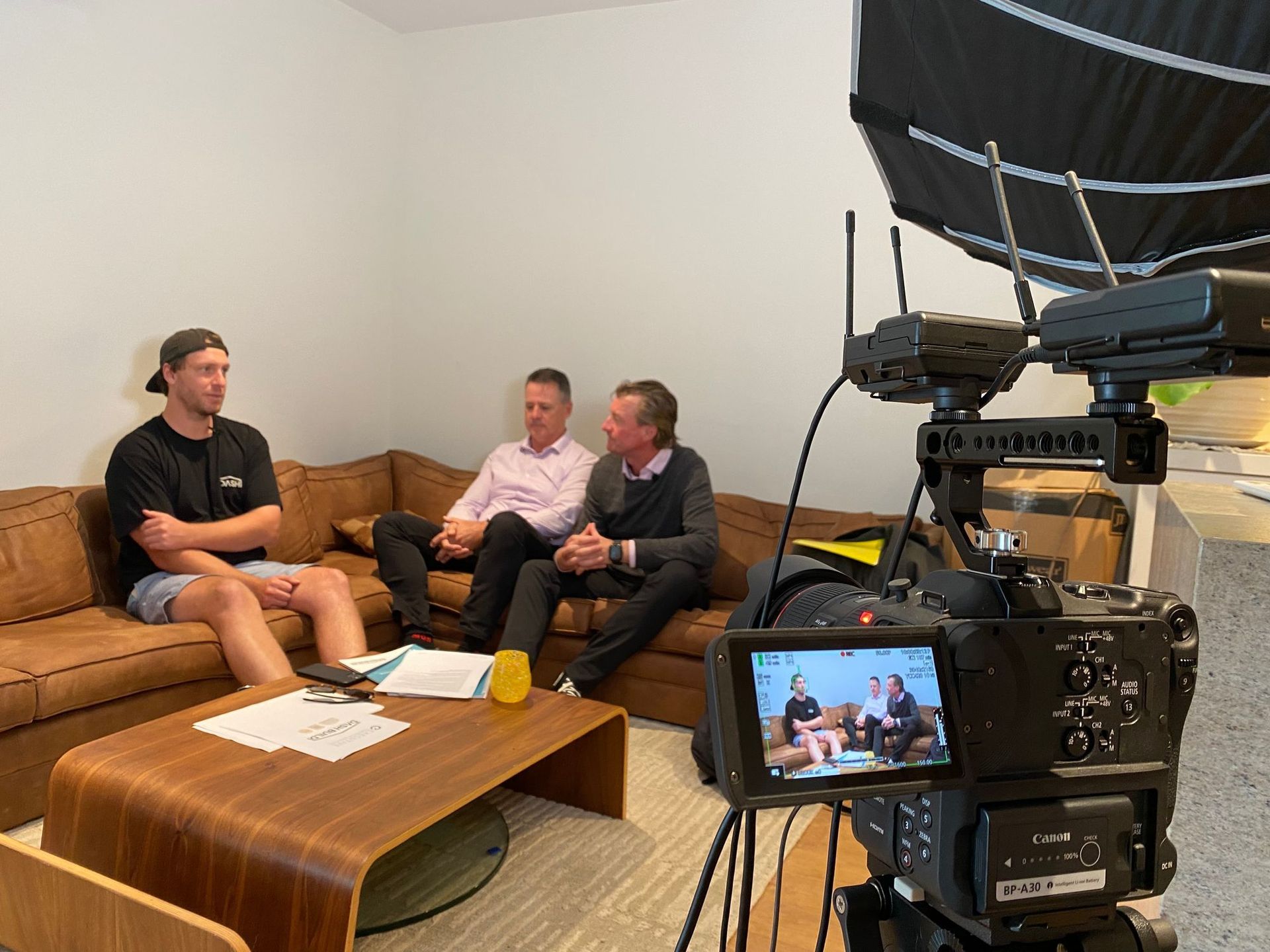 Three men on a brown couch in a room. One in shorts, two in business attire. Filming with a camera.