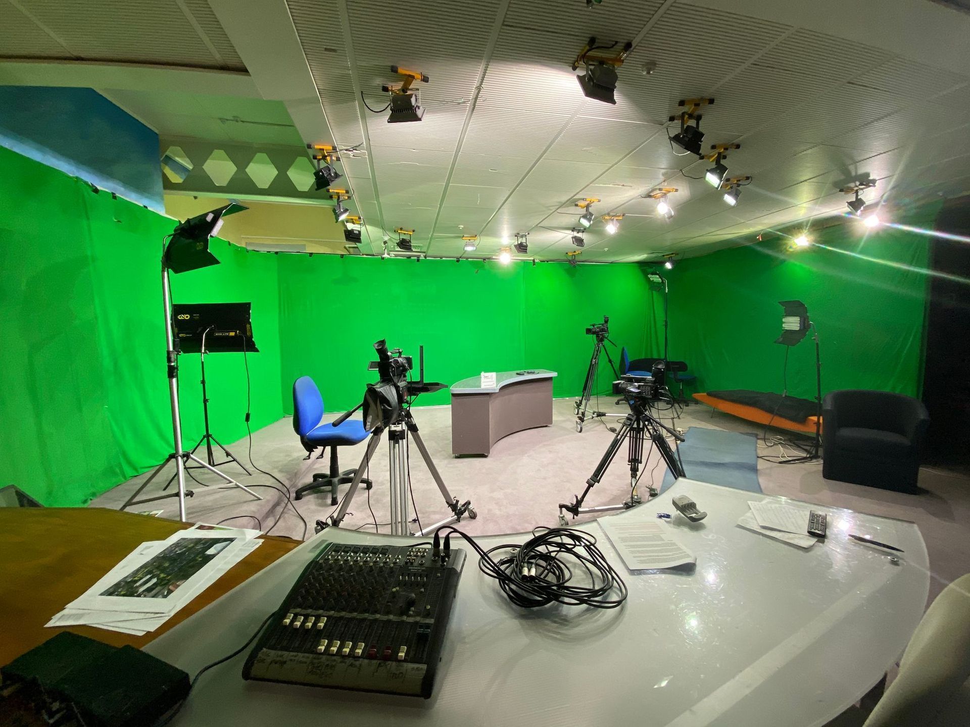Television studio with green screen, cameras on tripods, lights, control desk, and a desk with a chair.