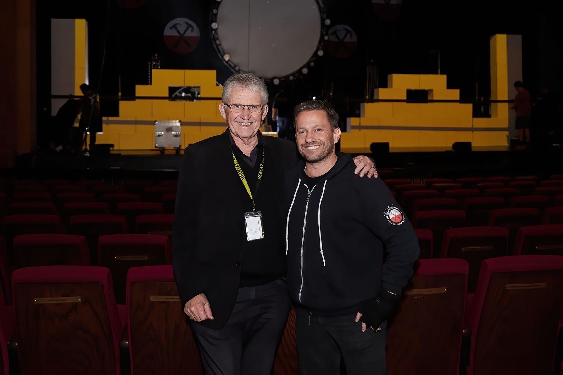 Two men stand in theater: One older in black suit, one younger in hoodie, arms around each other. Stage in background.