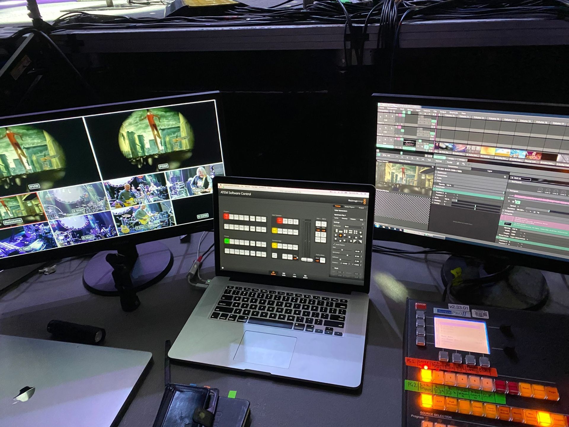 A media editing workspace with many monitors displaying video feeds and editing software. Laptop and control panel visible.