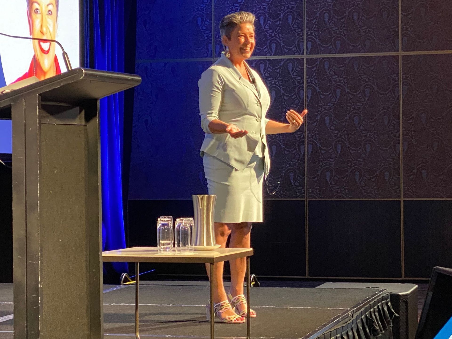 Woman in light suit speaking at a podium, gesturing with hands. Stage with screen, water glasses.