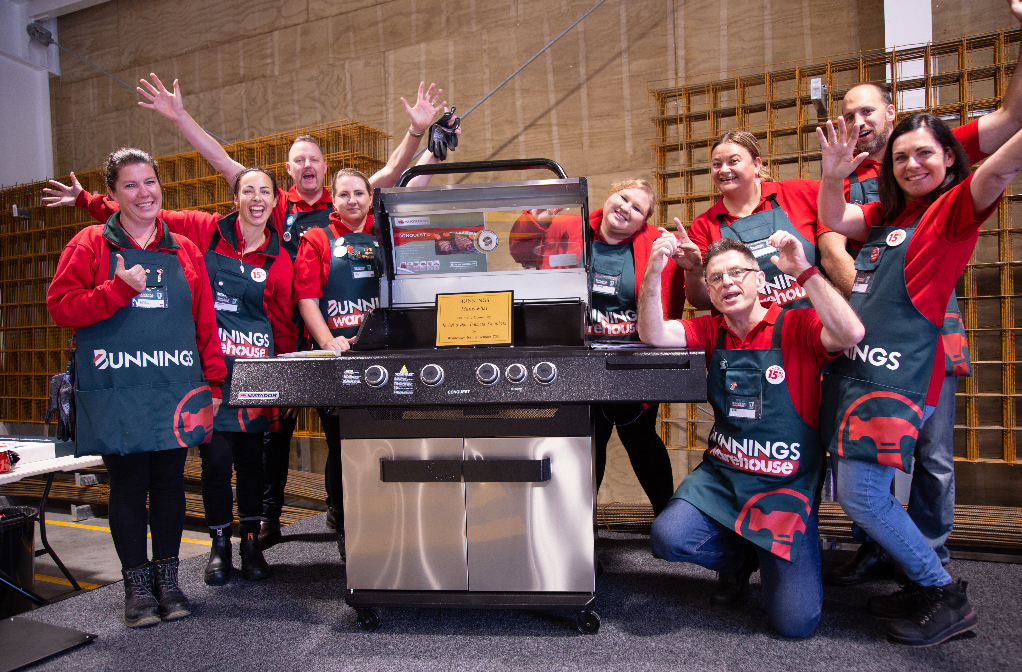 Group of people wearing red shirts and aprons, celebrating around a large barbecue grill.