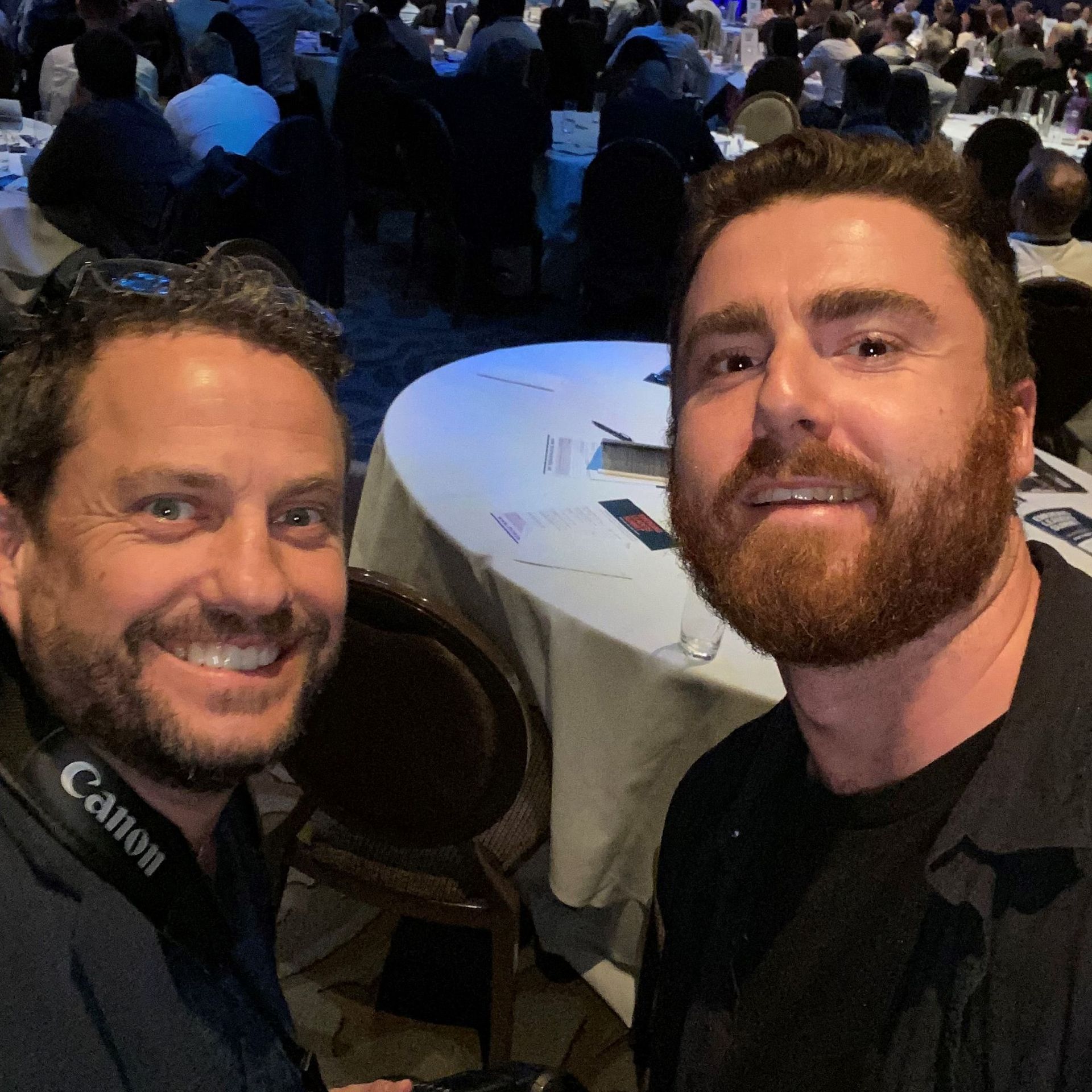 Two men smiling for a selfie at a formal event, with people and round tables in the background.
