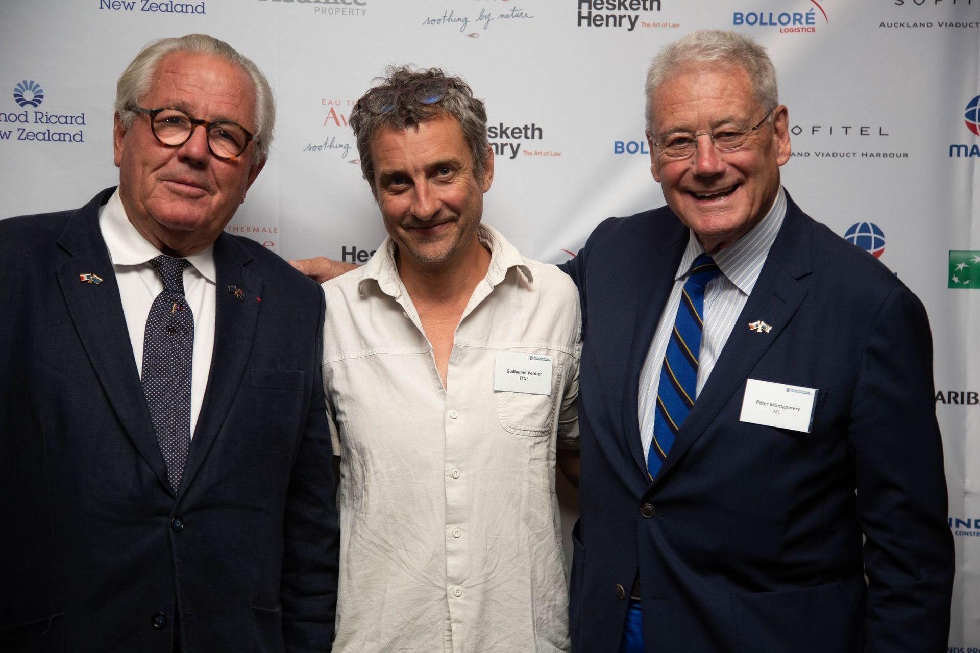 Three men in suits pose together at an event with a backdrop.