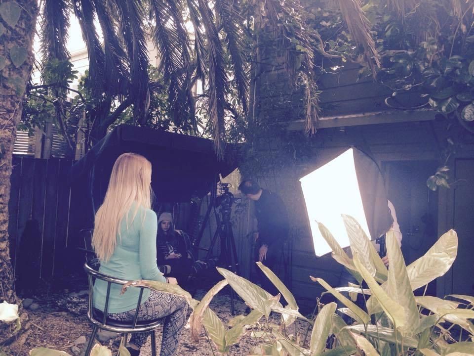 A woman with long blonde hair sits for an outdoor video shoot, with a camera and light.