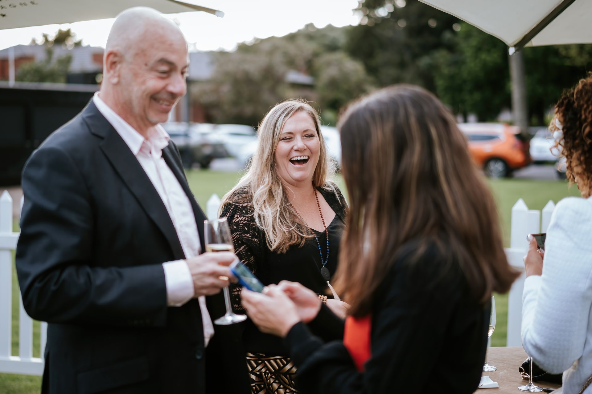 People at outdoor gathering, laughing. Man holding glass, woman in black dress, other woman facing them.