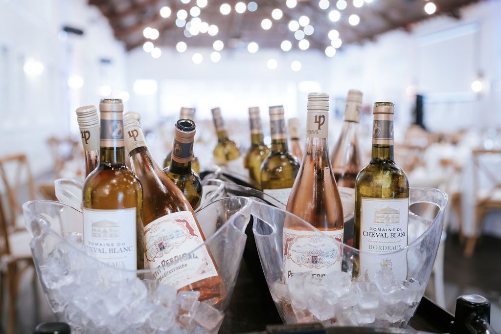 Wine bottles in ice buckets at an event. White interior, overhead string lights, and tables with chairs in the background.