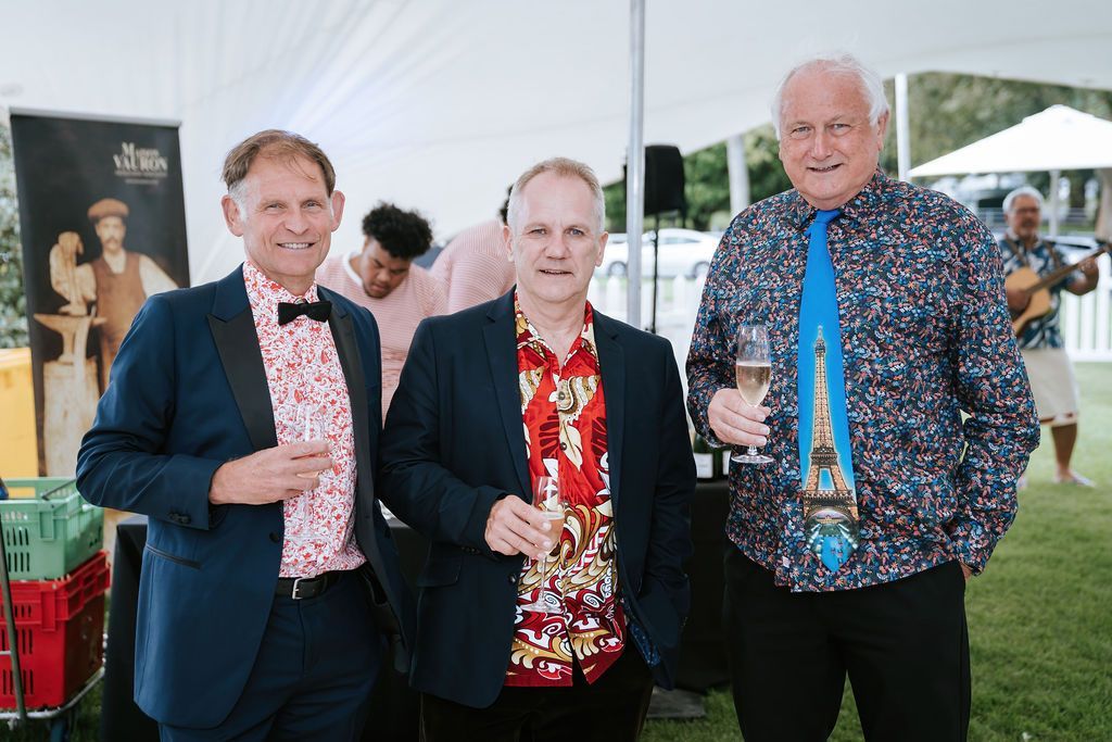 Three men in semi-formal attire, holding drinks, smiling outdoors. One man sports a whimsical tie.