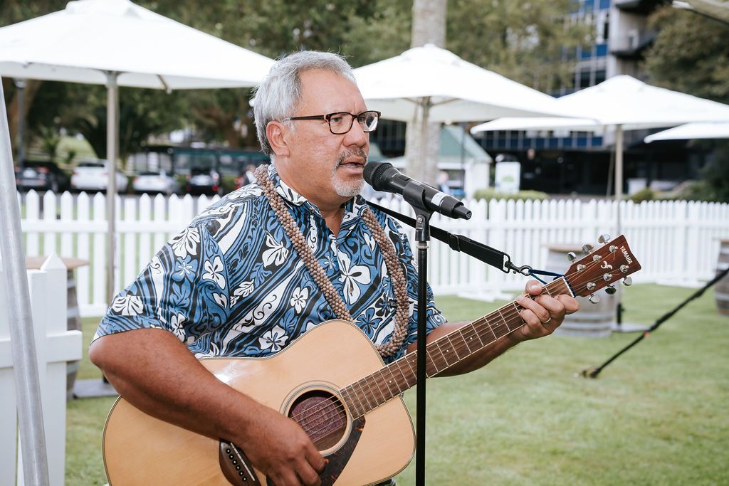 Man playing acoustic guitar, singing into a microphone outdoors. He's wearing glasses and a patterned shirt.