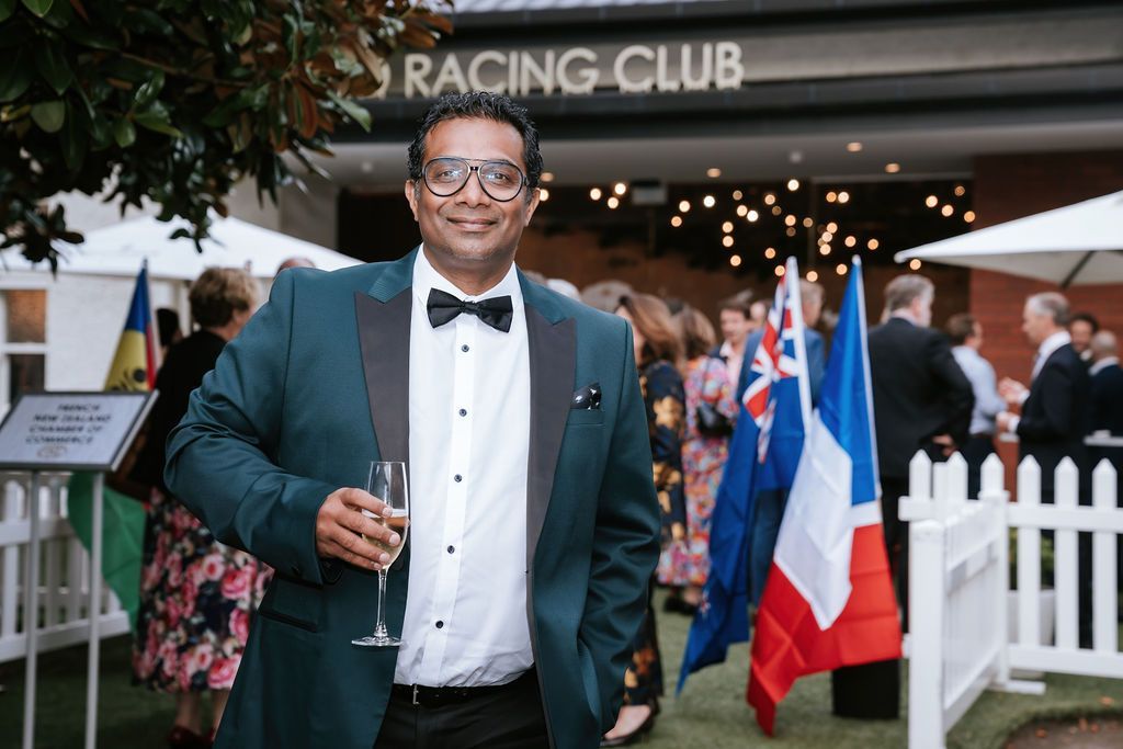Man in formal attire holding a champagne glass at a racing club, surrounded by flags and people.