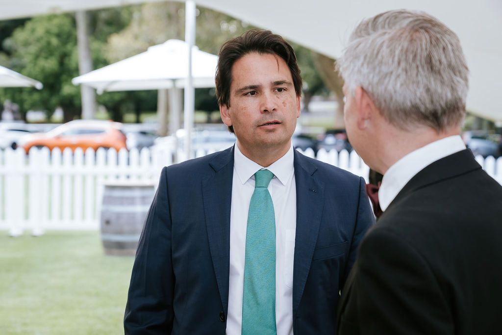 Man in a blue suit and teal tie talking to another man in a dark suit outdoors.