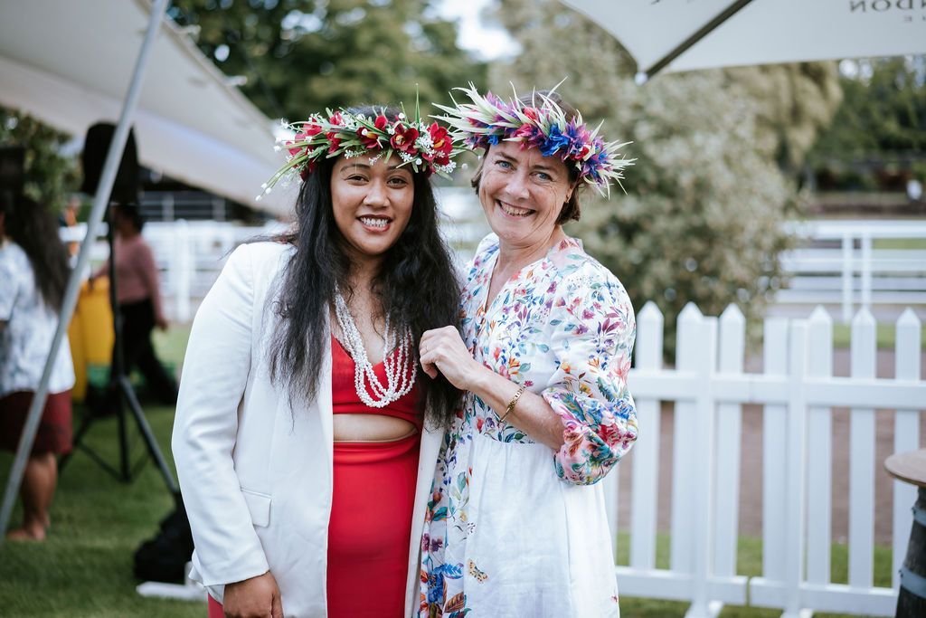 Two women wearing floral crowns smile outdoors, standing near a white picket fence.