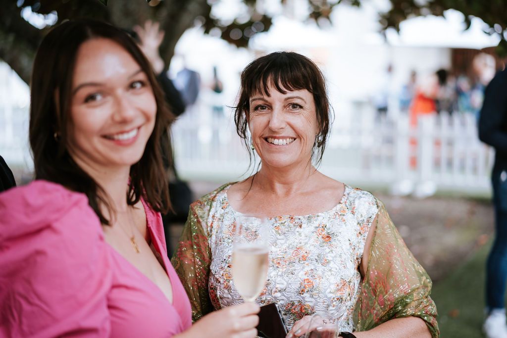 Two smiling women outdoors, one holding a glass. The woman on the left wears pink, the other floral.