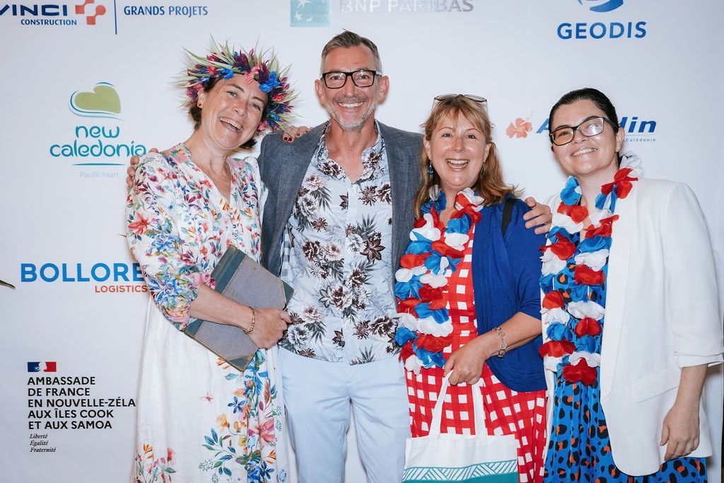 Four people smiling in front of a backdrop, two wearing leis and one with floral headpiece.