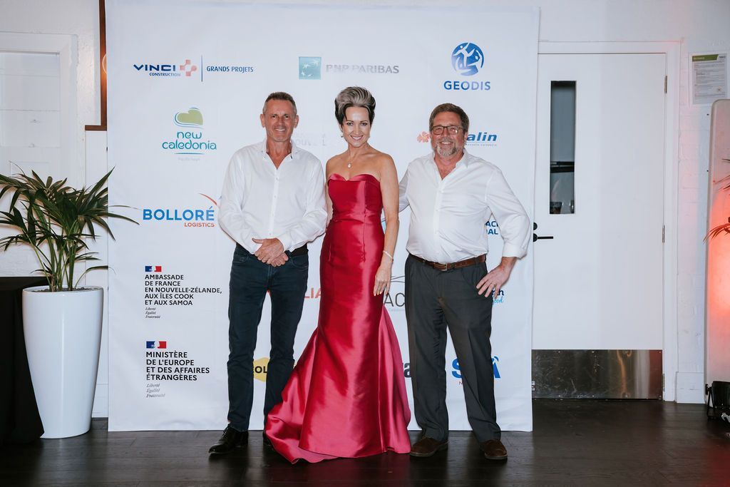 Three people posing in front of a backdrop at an event; woman in red gown stands between two men in white shirts.