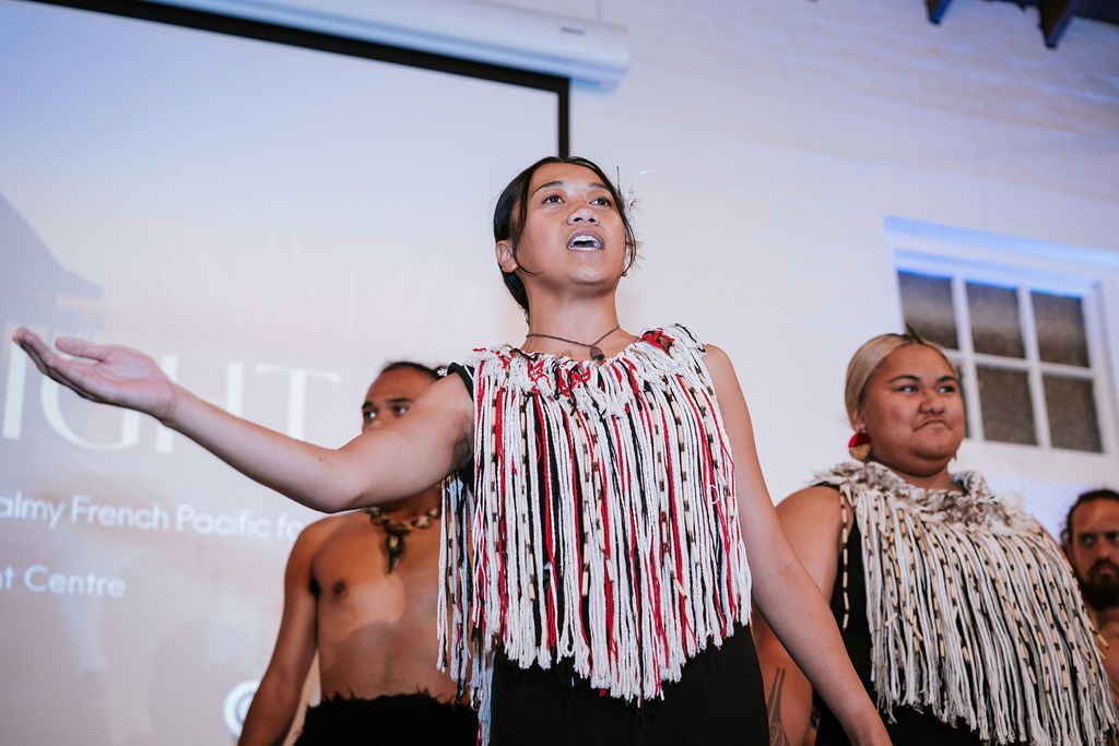 Woman performing, wearing traditional fringed garment, arms raised. Others stand behind. Projector screen in background.