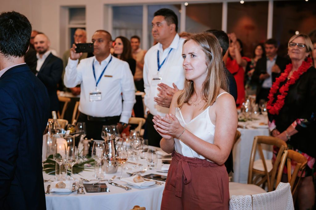 People clapping at a formal event, tables set with place settings, indoor setting.