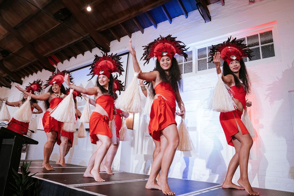 Dancers in red dresses and headdresses perform on a stage, holding white fans.