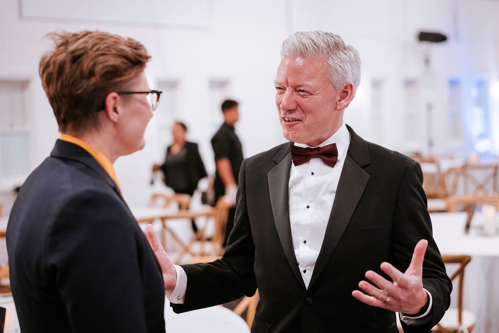 Man in tuxedo and bow tie gestures while speaking to person in glasses and blazer at an event.