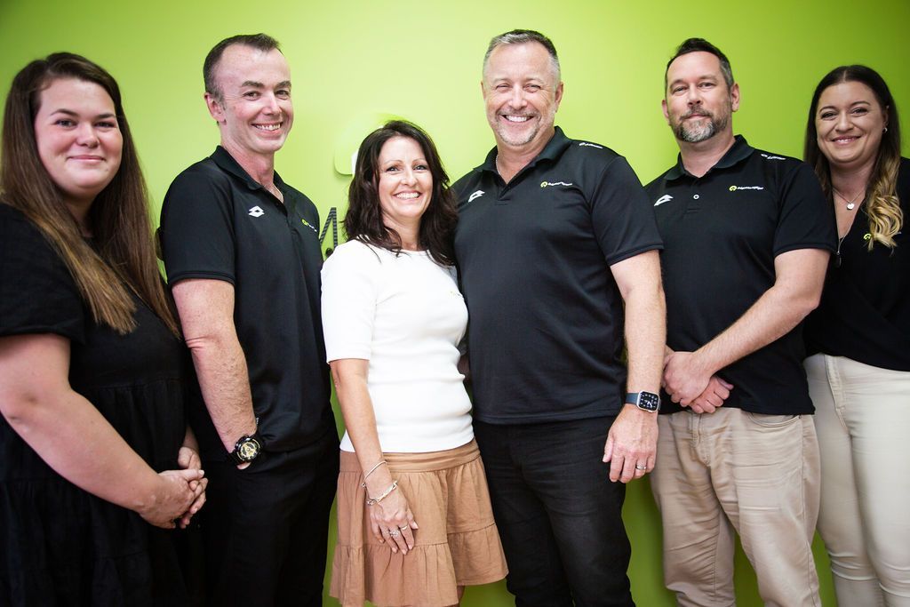 Six people standing and smiling in front of a green wall, wearing black and white clothing.