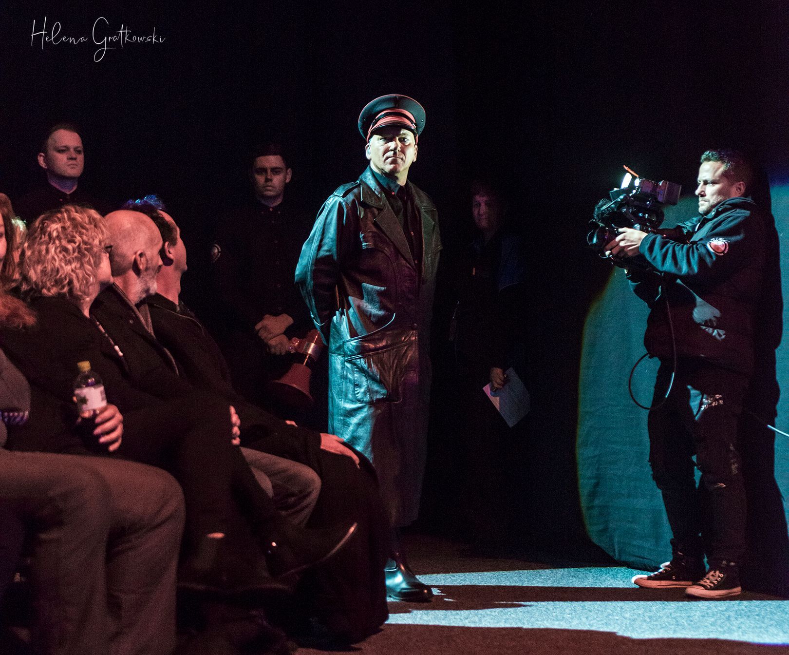 Man in military-style coat and cap, standing on stage. A cameraman films him while spectators watch.
