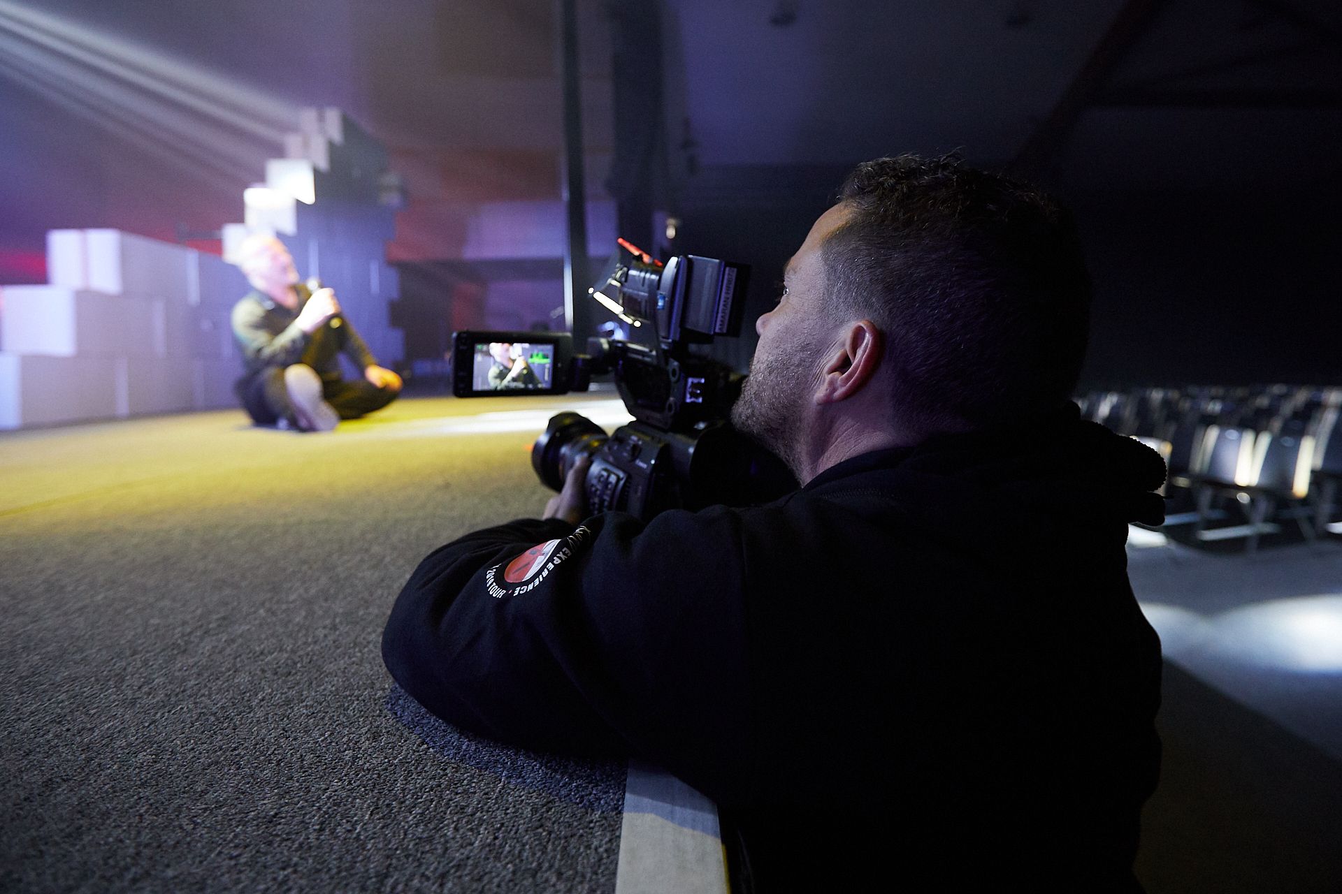 A person filming a seated individual with a camera in a studio setting with stairs and colorful lighting.