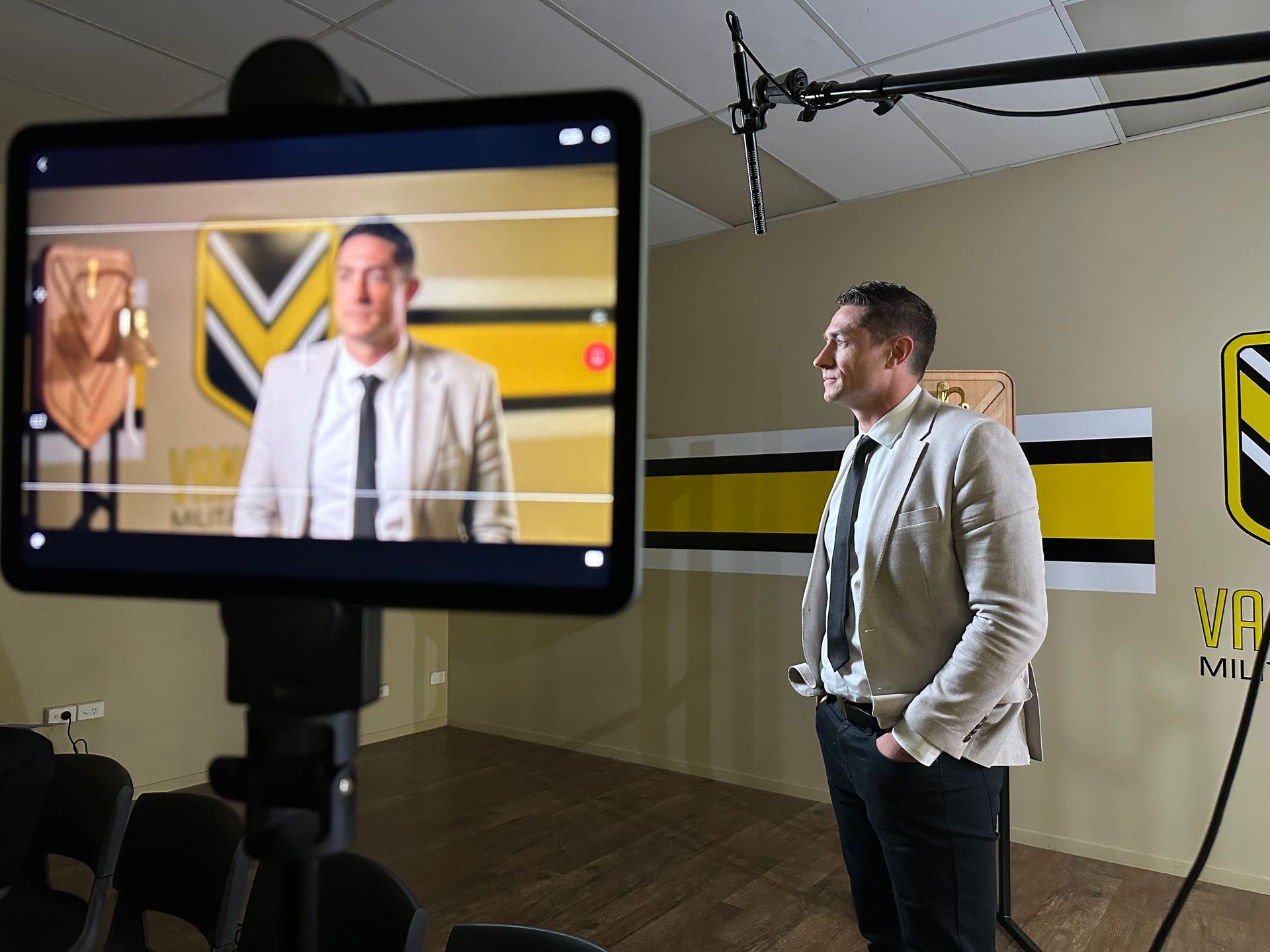 Man in suit stands beside a screen displaying a broadcast. Behind, a logo and yellow/black stripes decorate the wall.