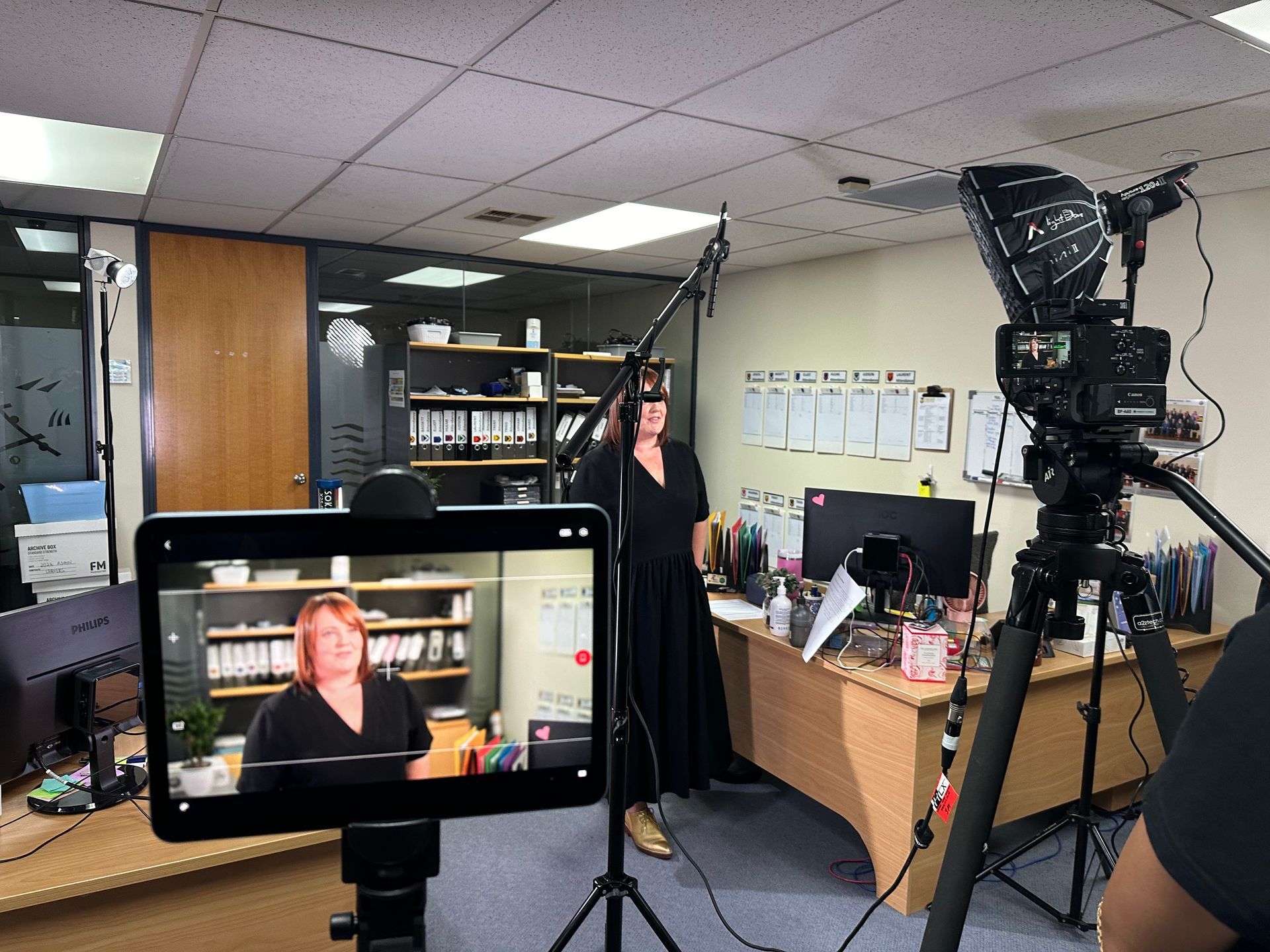 A woman in a black dress filming a video in an office, with cameras and equipment set up around her.