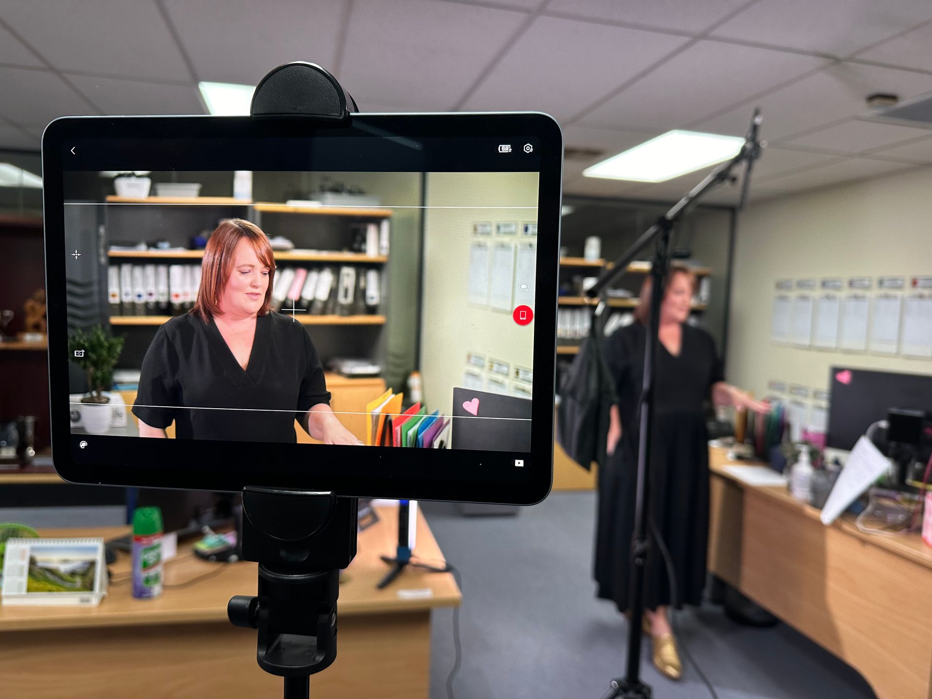 A woman in black dress filming a video in an office. Tablet on tripod displays the video feed.